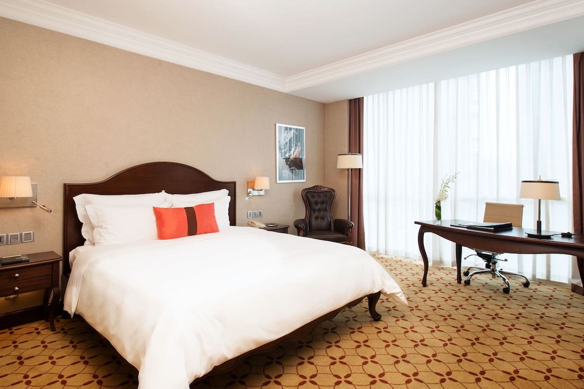 Una habitación de hotel con una cama grande, un escritorio, una silla y un ventanal con cortinas.