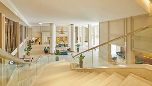 Un vestíbulo amplio con una escalera monumental, grandes columnas y una zona de estar con muebles modernos.
