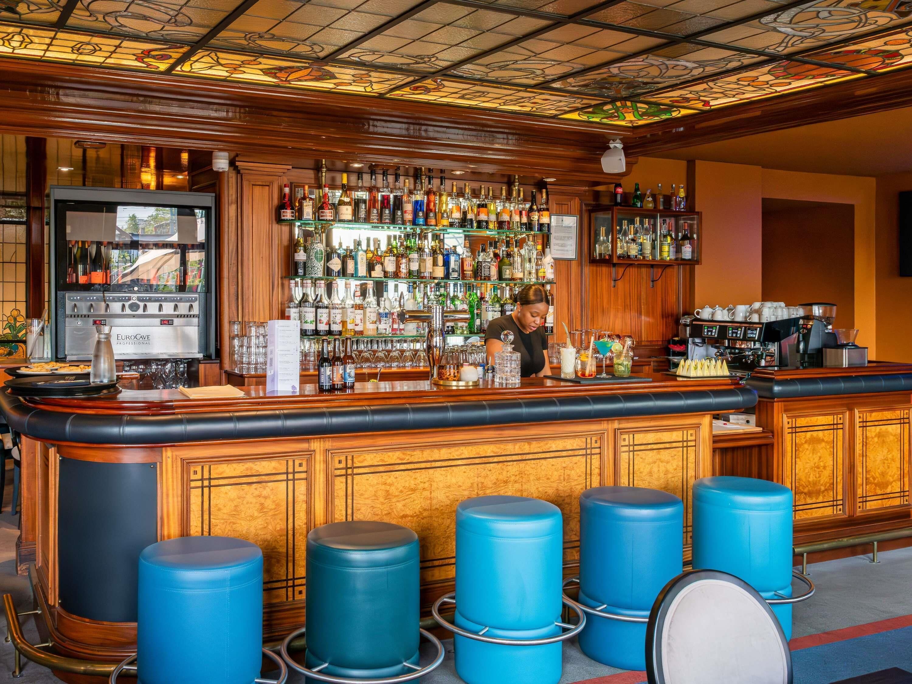 Un bar con mostrador de madera, varios taburetes y una amplia variedad de botellas y vasos.