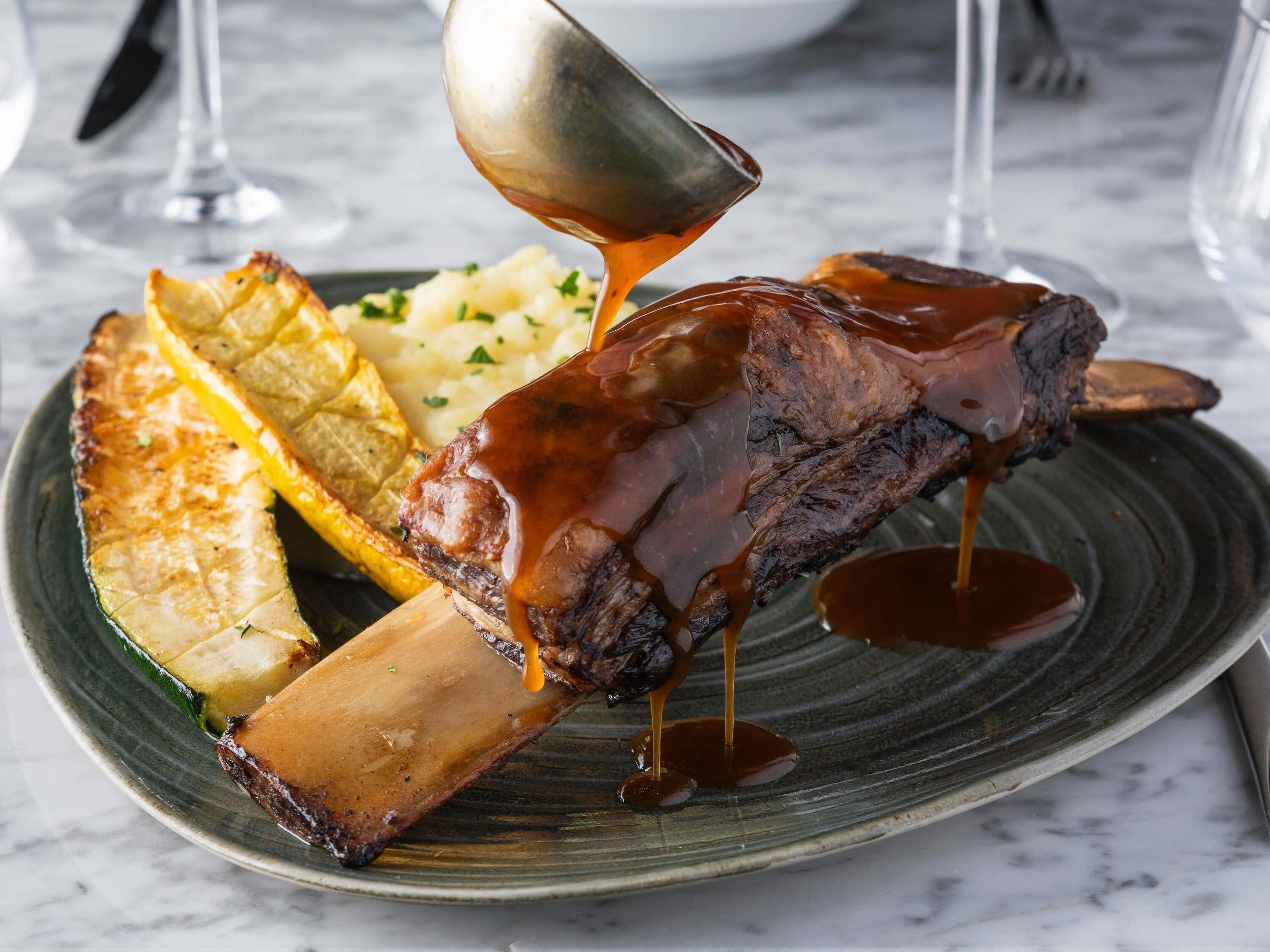 Une assiette contenant un os rôti, de la purée de pommes de terre et des légumes rôtis.