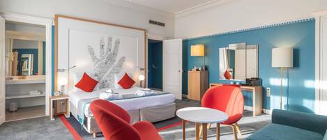 A hotel room with a bed, two red chairs, a teal sofa, a small white table, and a blue wall.