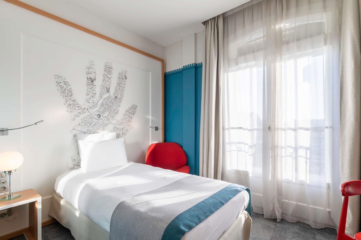 Une chambre d’hôtel avec un lit, un fauteuil rouge, une table de chevet, une lampe et une grande fenêtre avec des rideaux légers.