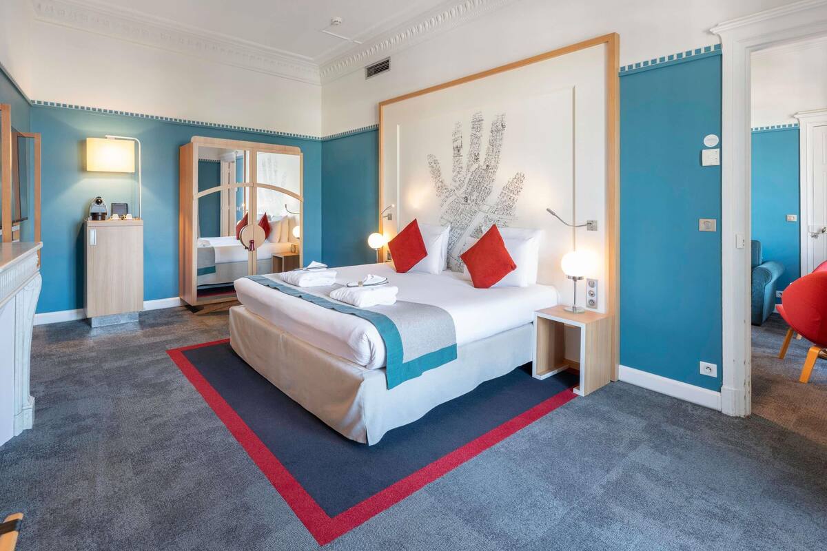 Une chambre d’hôtel avec un grand lit, une table de chevet avec une lampe, un miroir et un mur orné d’un motif décoratif.