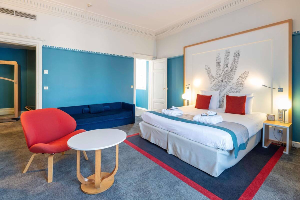 Une chambre d’hôtel avec un lit, une chaise rouge, une table ronde et un canapé bleu.