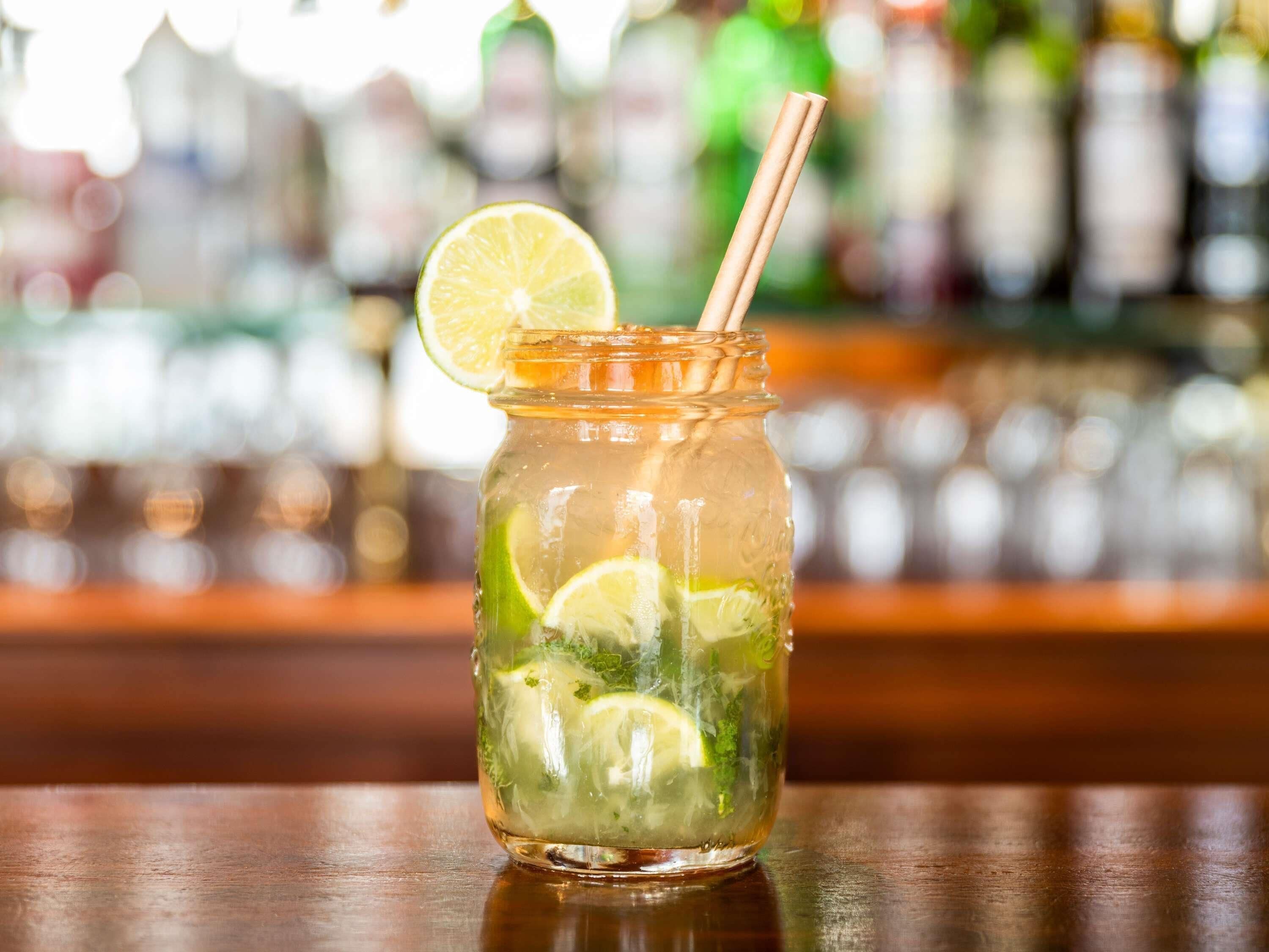 Un frasco de vidrio con una rodaja de lima y una pajita, conteniendo una bebida con hielo y hierbas, sobre una superficie de madera con un fondo desenfocado de botellas.