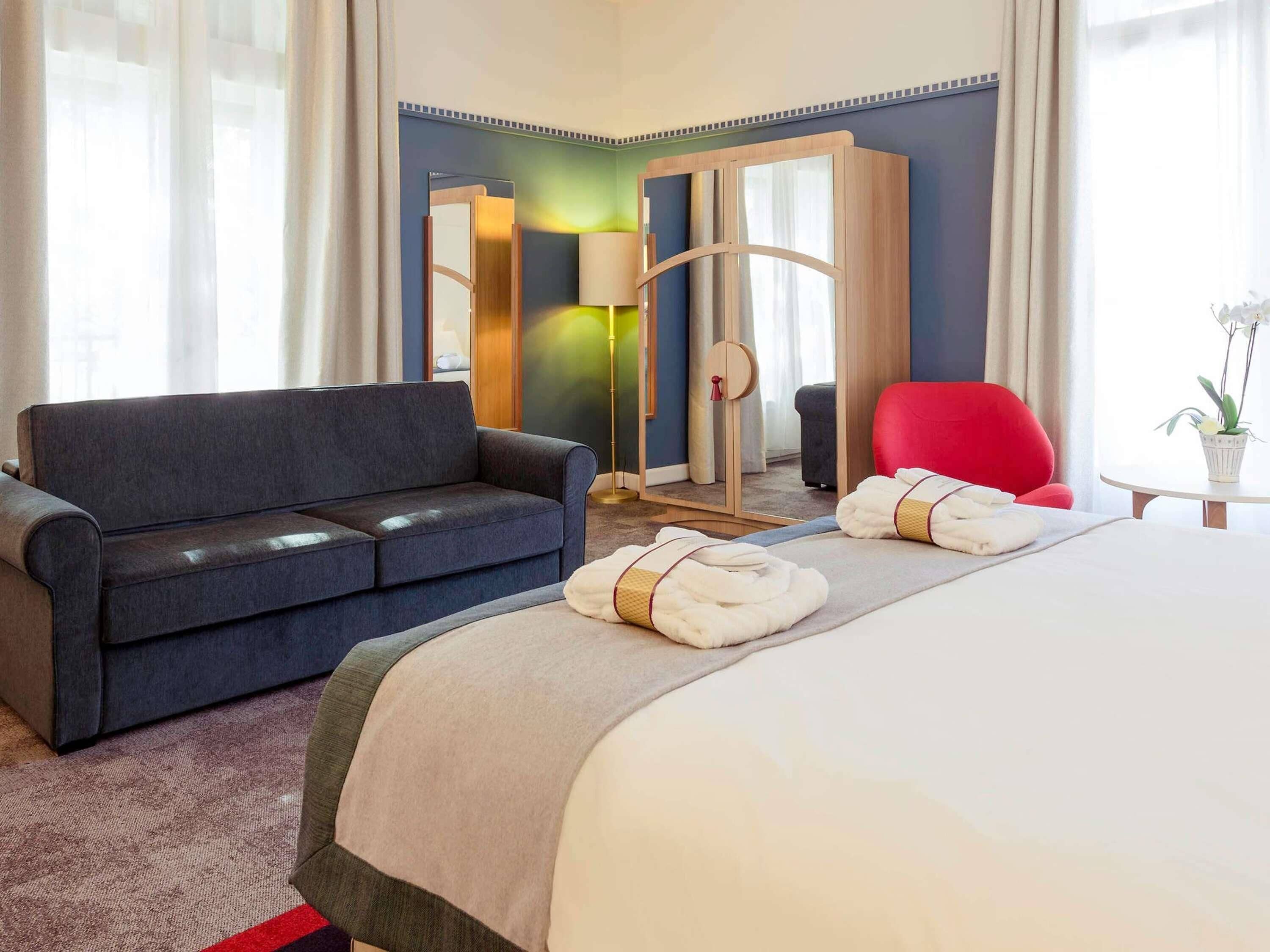 Ein Hotelzimmer mit Bett, einer Couch, einem roten Sessel, einem kleinen Tisch mit Pflanze und einem Spiegel.