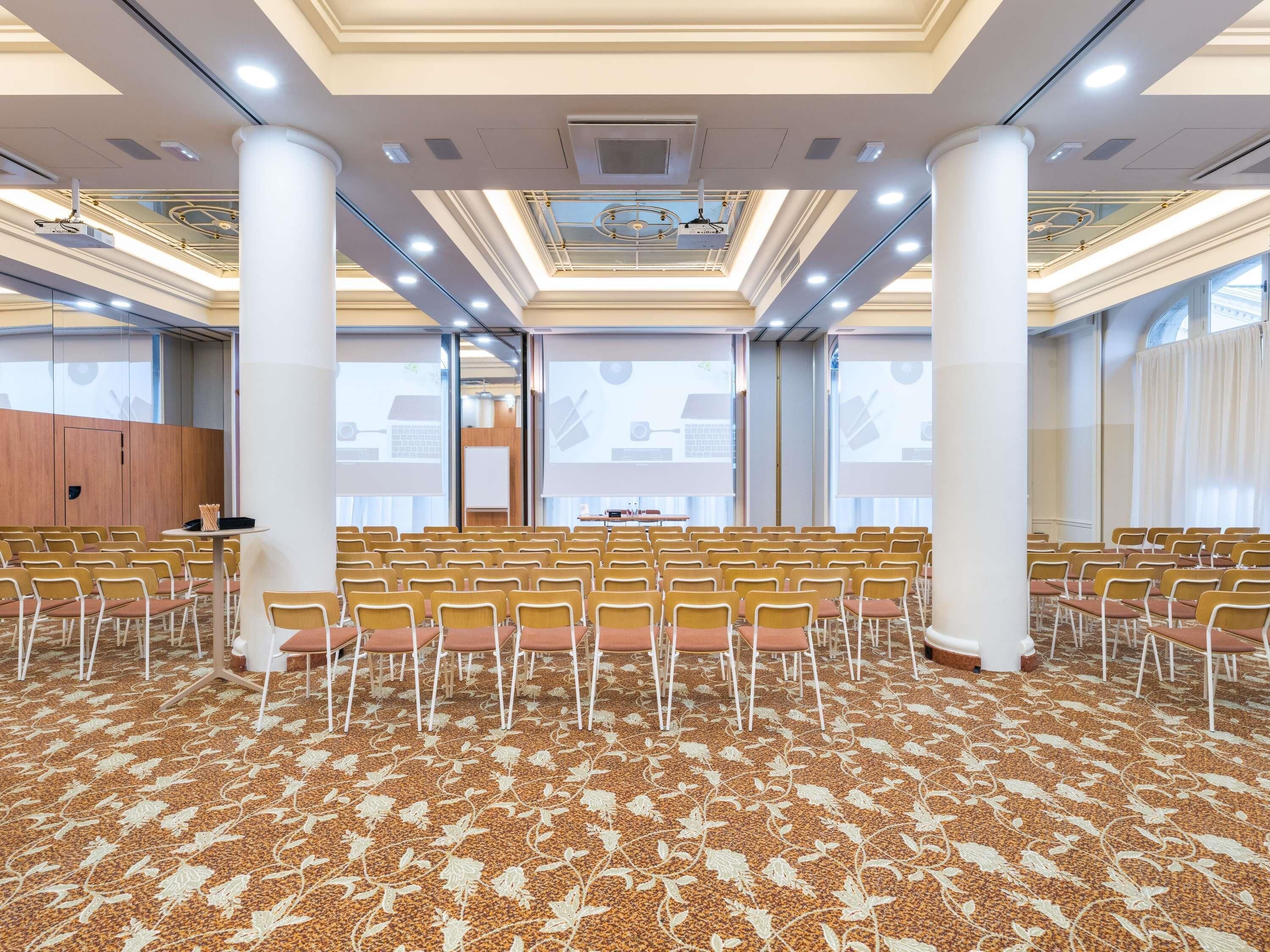 Un salón de conferencias con filas de sillas, un atril y una pantalla grande.