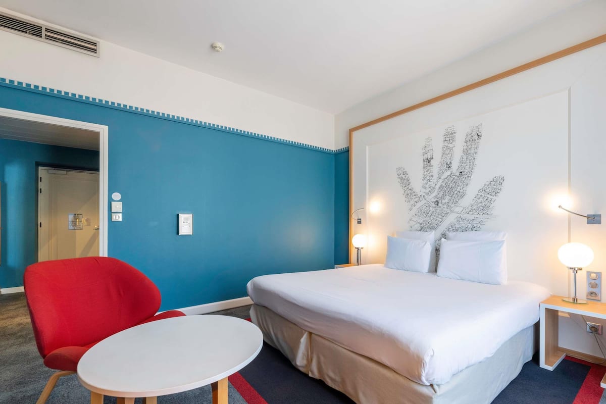 Une chambre d’hôtel avec un lit, une chaise rouge, une petite table et un mur orné d’une grande œuvre d’art abstraite.