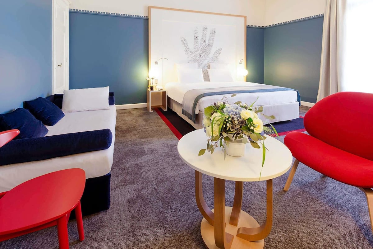 Une chambre d’hôtel avec un lit, un canapé, un fauteuil rouge, une petite table sur laquelle est posé un vase de fleurs, et une œuvre d’art murale.