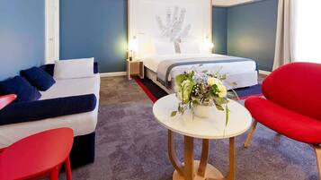 Habitación de hotel con cama, sofá, una silla roja, una mesita con un jarrón de flores y una obra de arte en la pared.