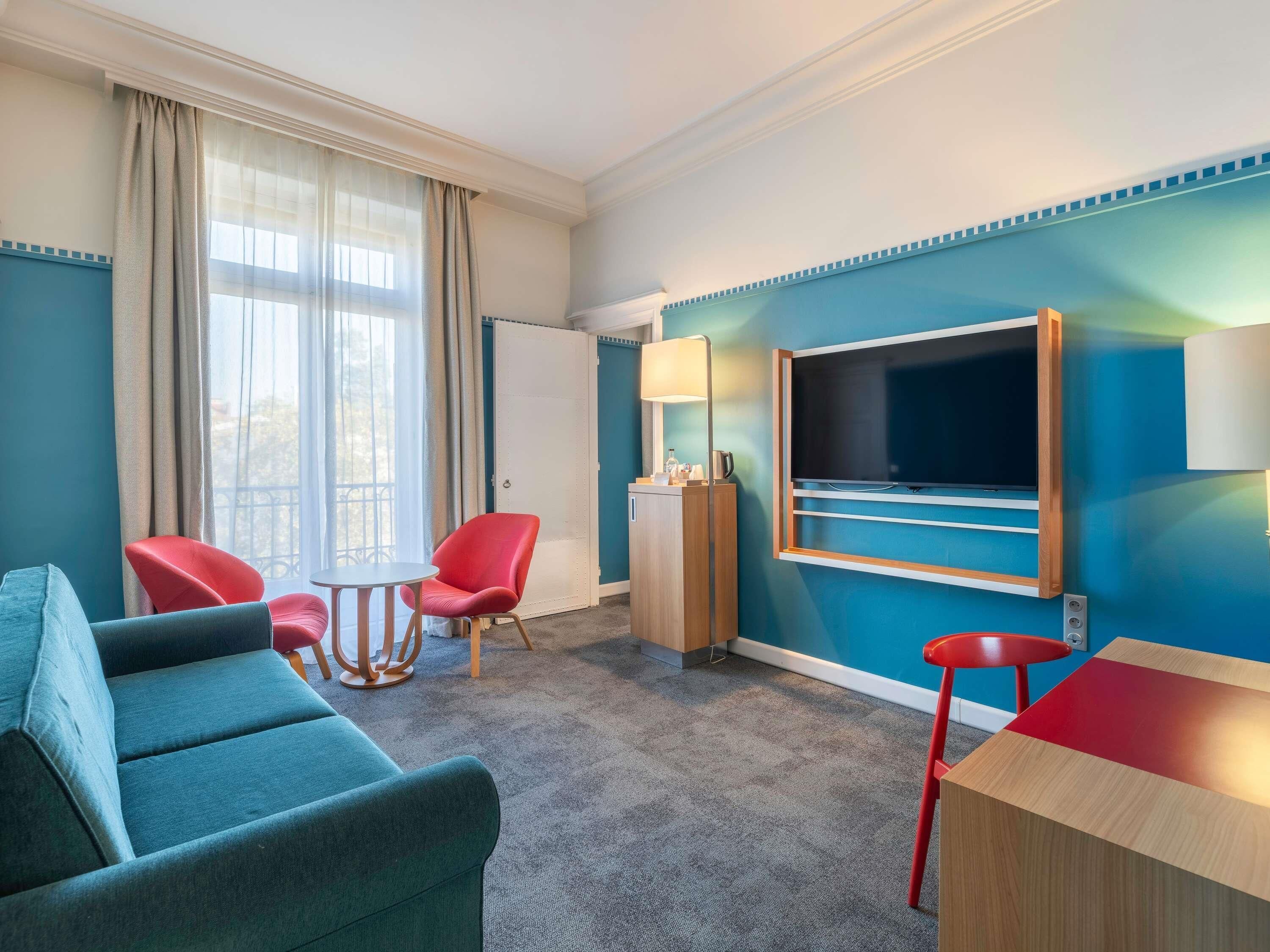 Une chambre d’hôtel avec un mur de couleur turquoise, un grand téléviseur à écran plat fixé au mur, un fauteuil rouge, un canapé turquoise et une petite table ronde.