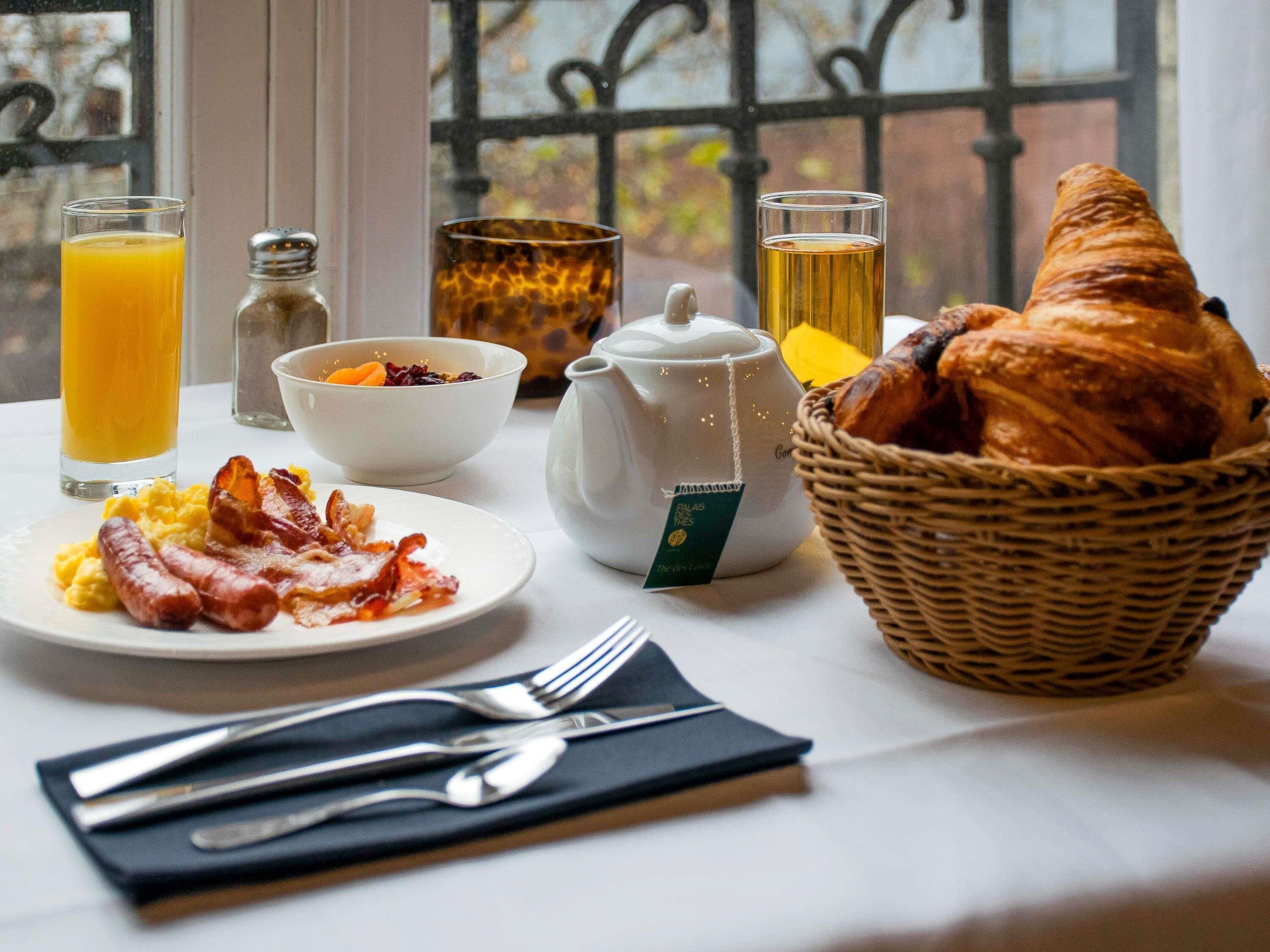 Une table dressée pour le petit-déjeuner avec un panier de croissants, des œufs brouillés, du bacon, un saladier de fruits, une théière, ainsi que des verres de jus d’orange et de thé.