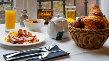 Un desayuno con una cesta de cruasanes, huevos revueltos, tocino, un tazón de fruta, un hervidor de agua y vasos de jugo de naranja y té.