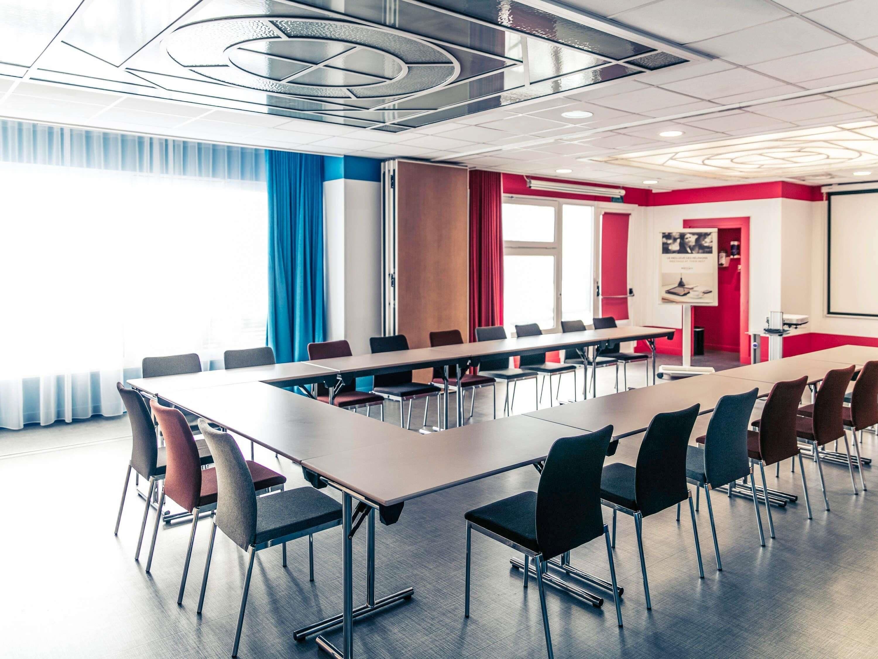 Uma sala de conferências com uma mesa comprida, cadeiras, um projetor e um amplo janelão com cortinas.