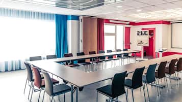 Una sala de conferencias con una larga mesa, sillas, una pantalla de proyector y un ventanal con cortinas.