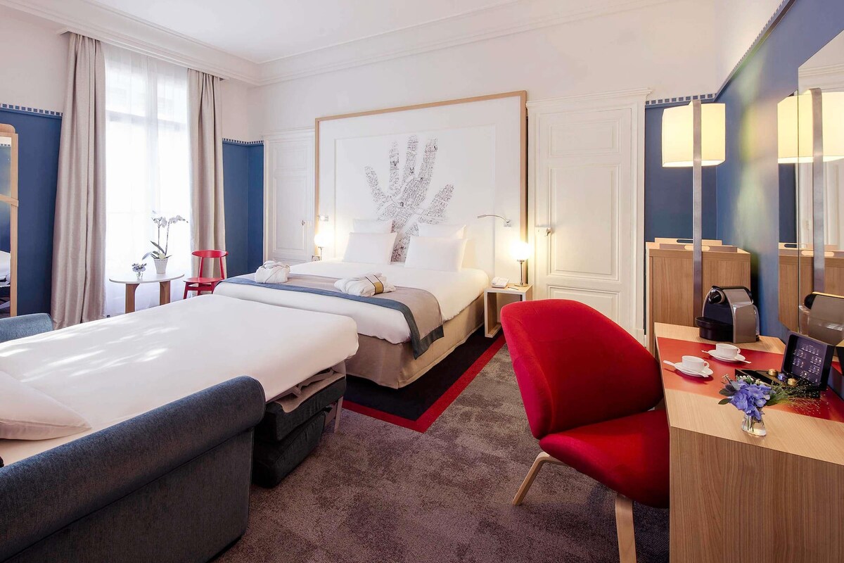 A hotel room with two beds, a desk with a coffee machine, a red chair, and a large framed artwork on the wall.