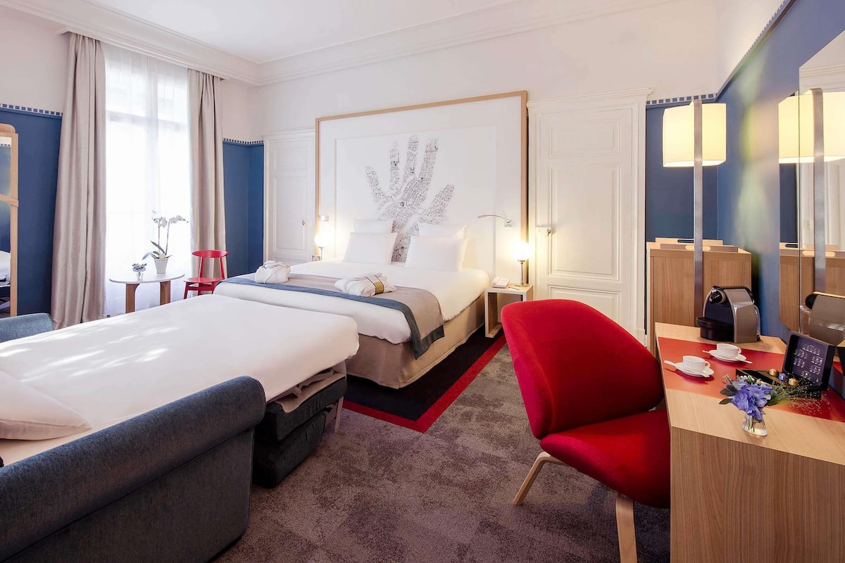 Hotellihuone, jossa on kaksi sänkyä, työpöytä, jossa on kahvinkeitin, punainen tuoli ja suuri kehystetty taideteos seinällä.