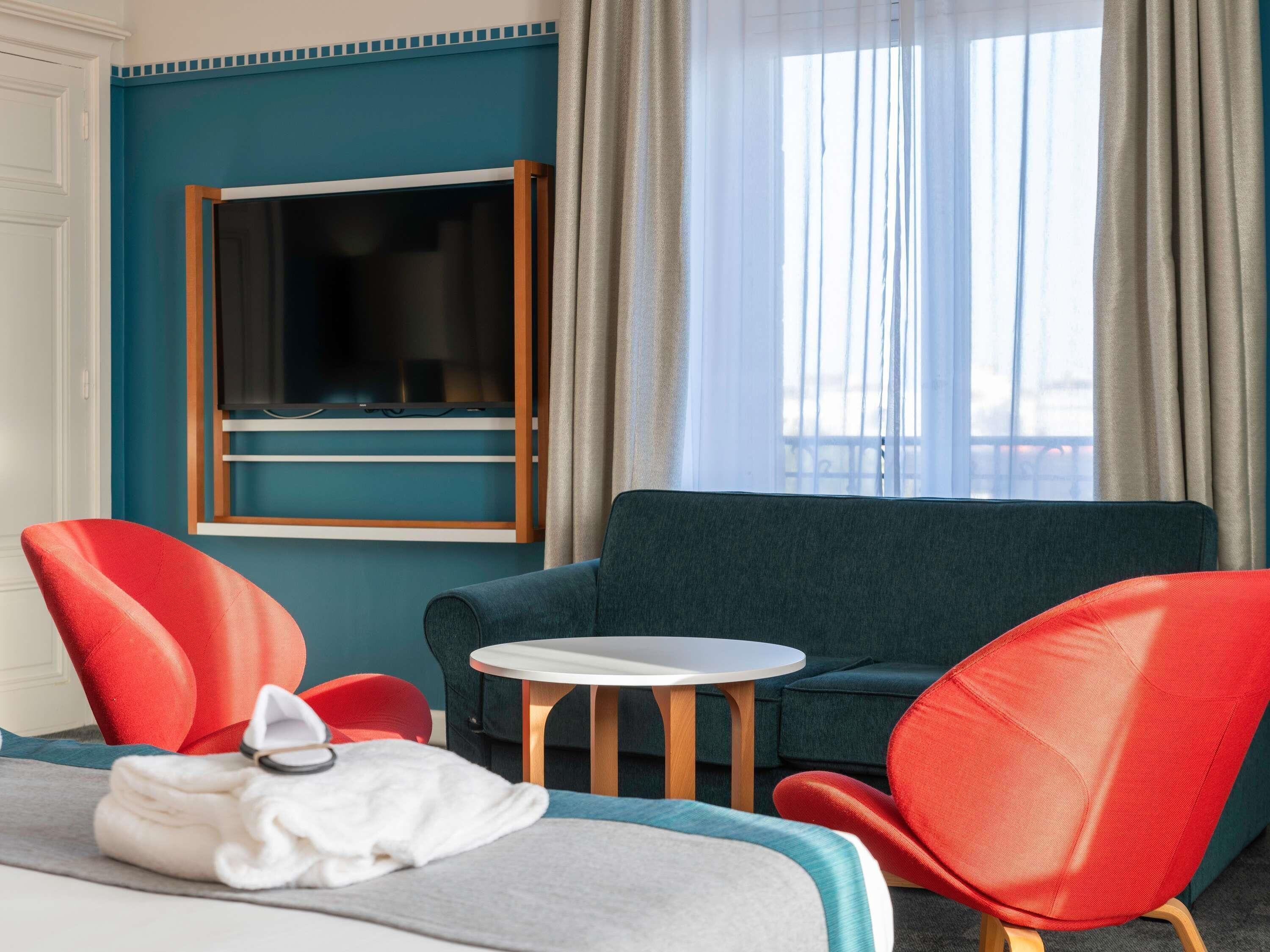 A hotel room with a bed, a blue sofa, two red chairs, a small white table, and a TV on a stand.