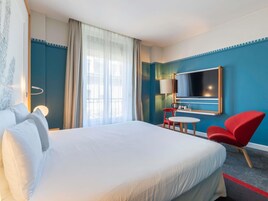 Hotellihuone, jossa on suuri sänky, työpöytä tuolilla, televisio ja ikkuna, jossa on verhot.