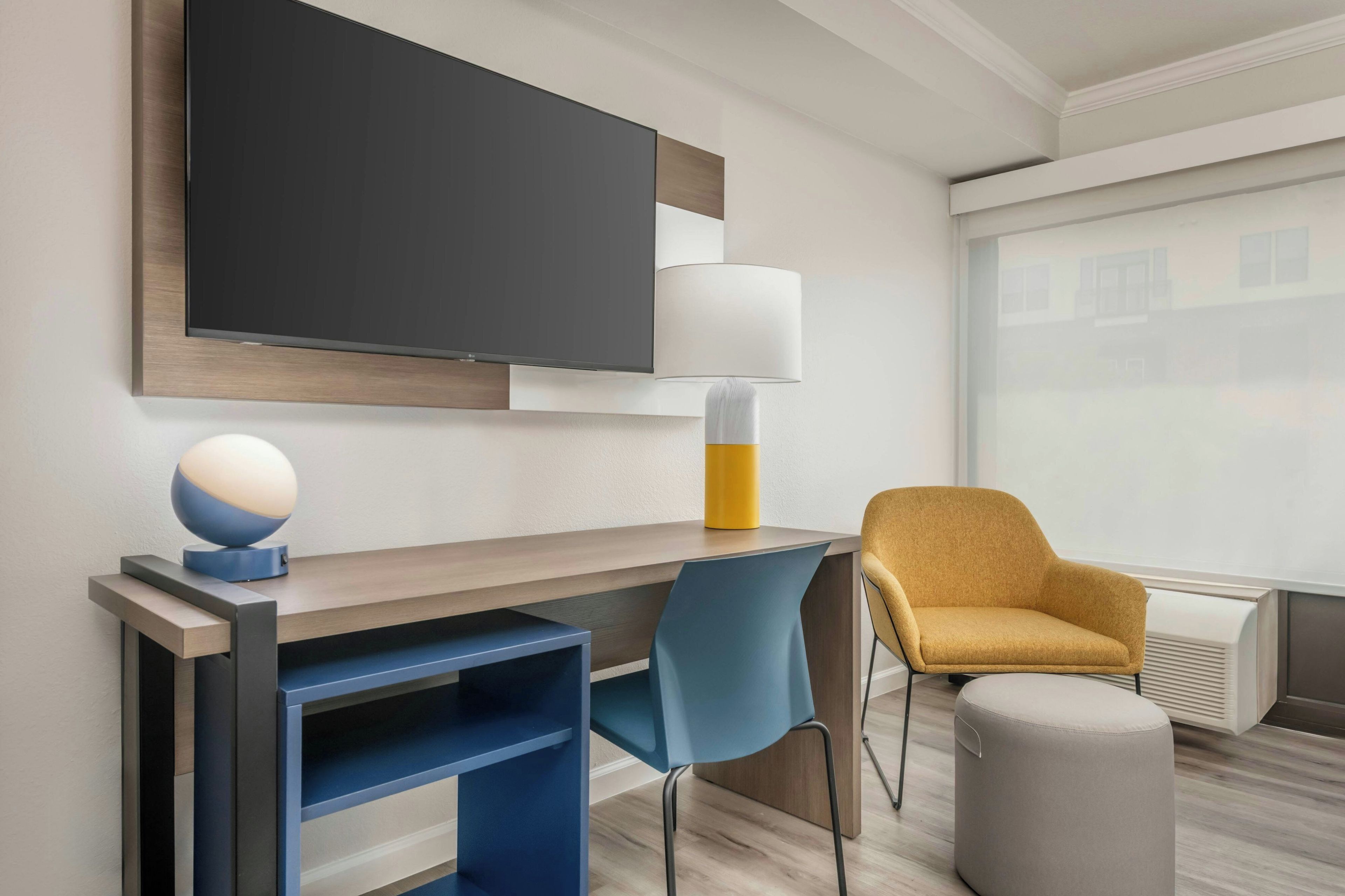 Quarto de hotel moderno com TV de tela plana, uma escrivaninha com luminária, uma cadeira azul, uma poltrona bege-mostarda e uma janela grande.