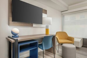 A modern hotel room with a flat-screen TV, a desk with a lamp, a blue chair, a mustard-colored armchair, and a large window. - City Express by Marriott I-10 West Energy Corridor (Houston)