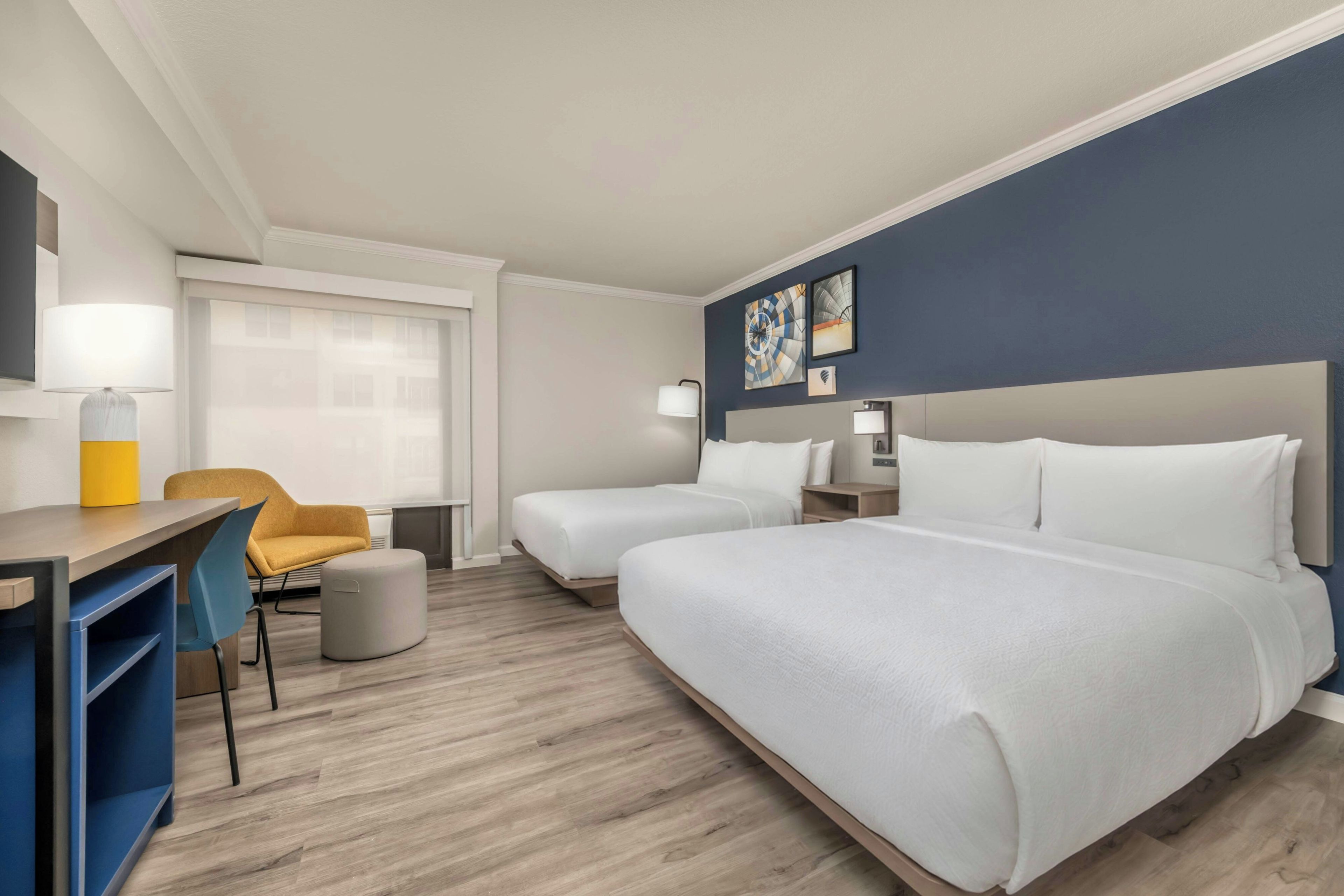 A hotel room with a desk, two beds, a chair, and artwork on the wall.
