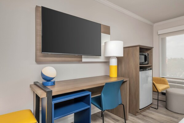 A hotel room with a wooden desk, a blue chair, a yellow ottoman, a television, a lamp, and a microwave. - City Express by Marriott I-10 West Energy Corridor (Houston)