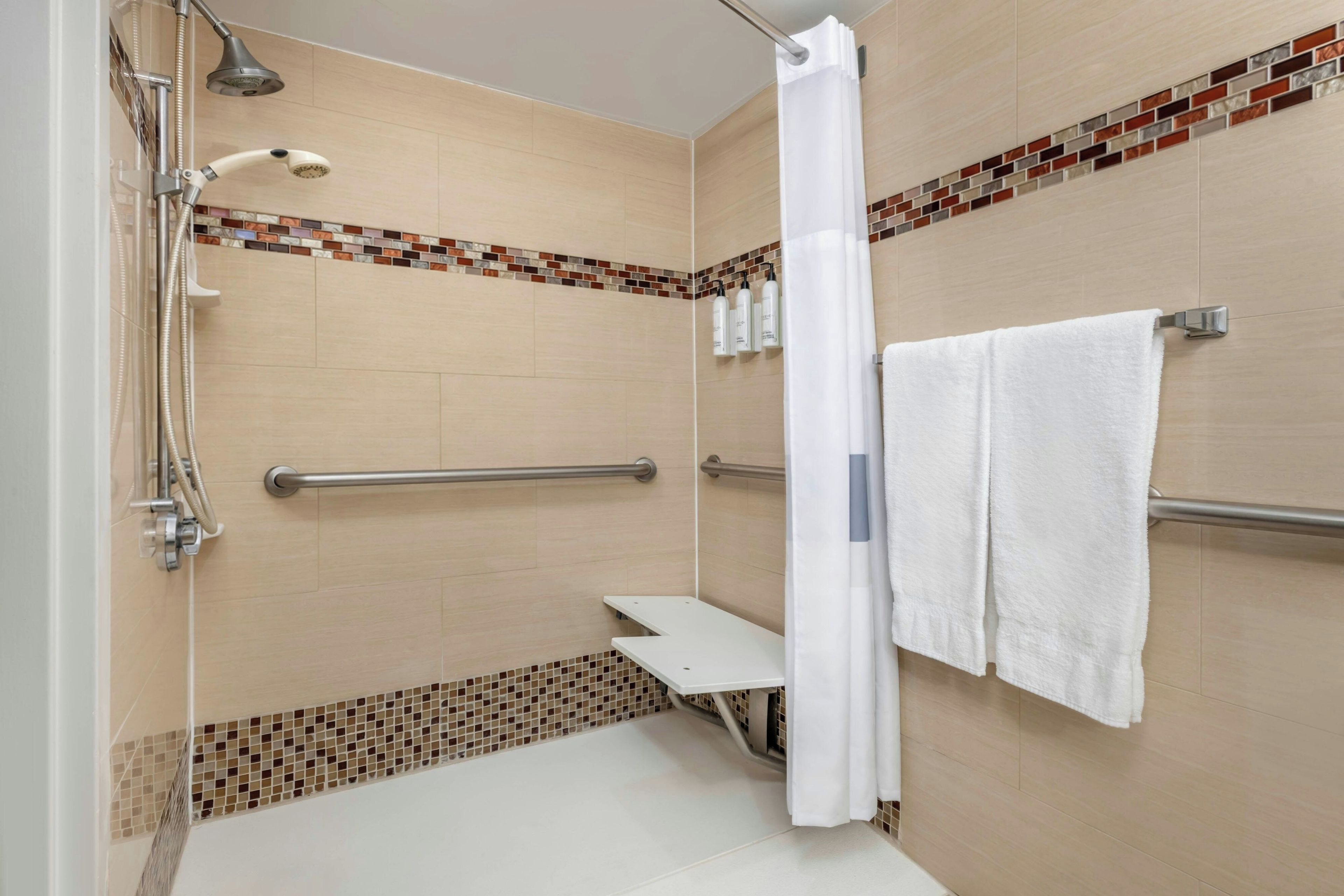 Zona de ducha con mampara de vidrio, cabezal de ducha manual, rociador de mano, barra de apoyo y toallero blanco.