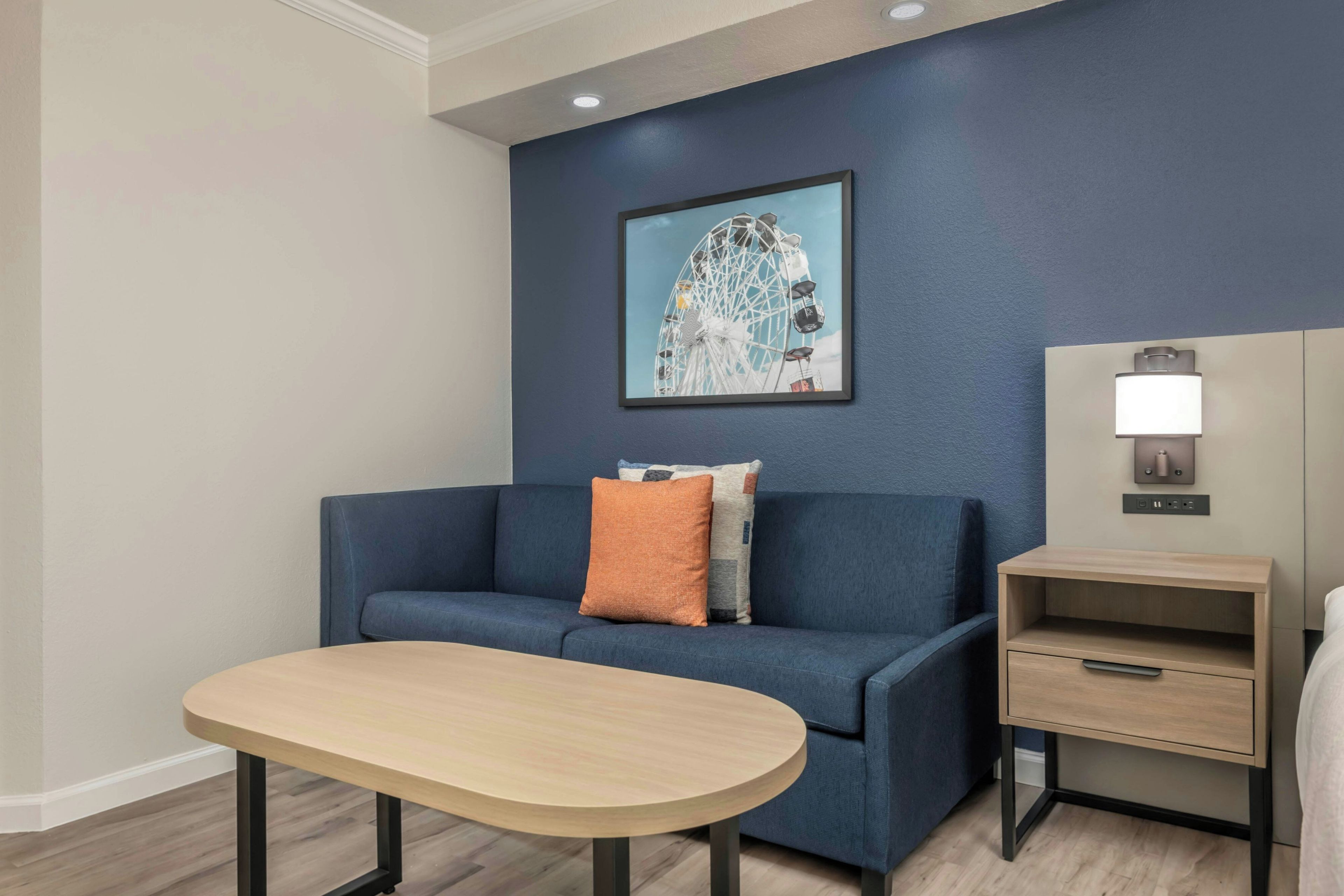 A modern room with a blue sofa, a small table, a nightstand with a lamp, and a framed picture on the wall.