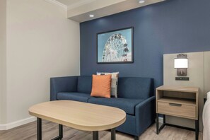 Una habitación moderna con un sofá azul, una mesita, una mesita de noche con lámpara y un cuadro en la pared.