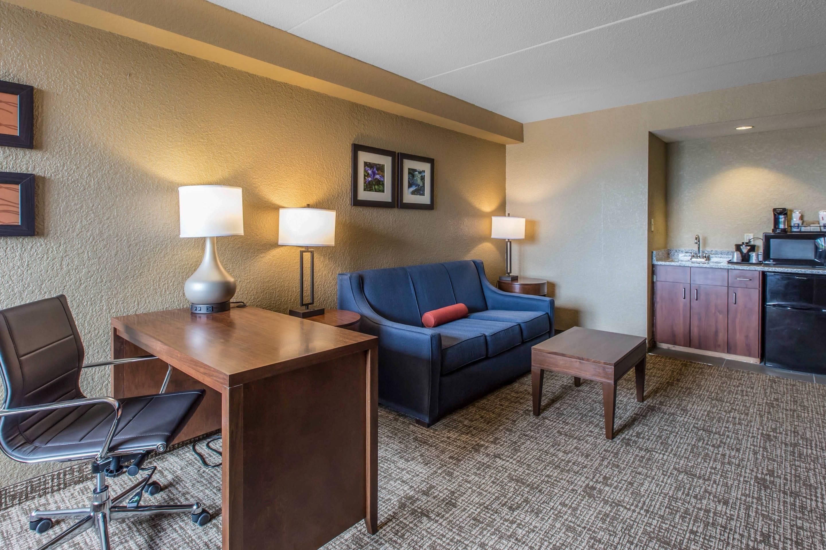 A hotel room with a desk, chair, sofa, and kitchenette.