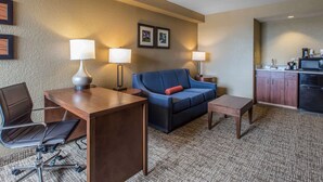 Une chambre d’hôtel comprenant un canapé, un bureau avec une chaise, une petite table, un coin cuisine et deux lampes fixées au mur.