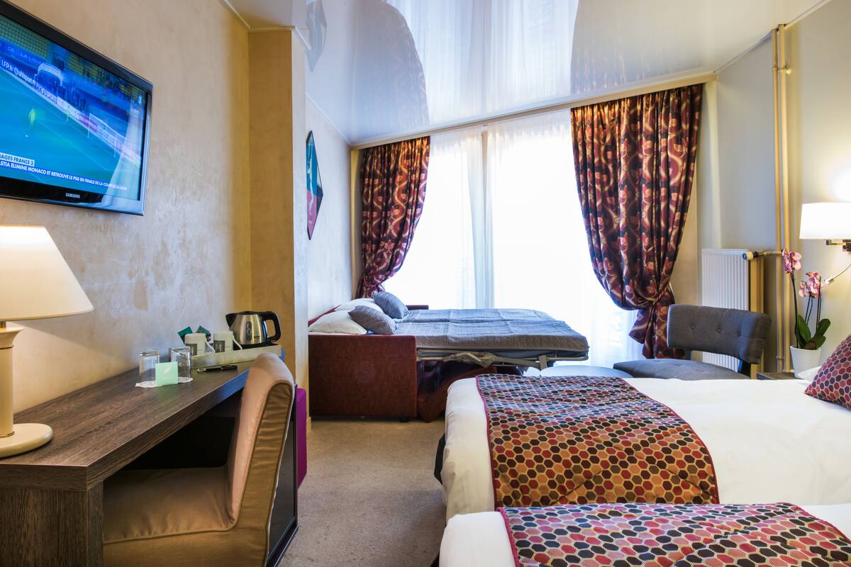 Una habitación de hotel con cama, escritorio, silla, televisión y ventana con cortinas.