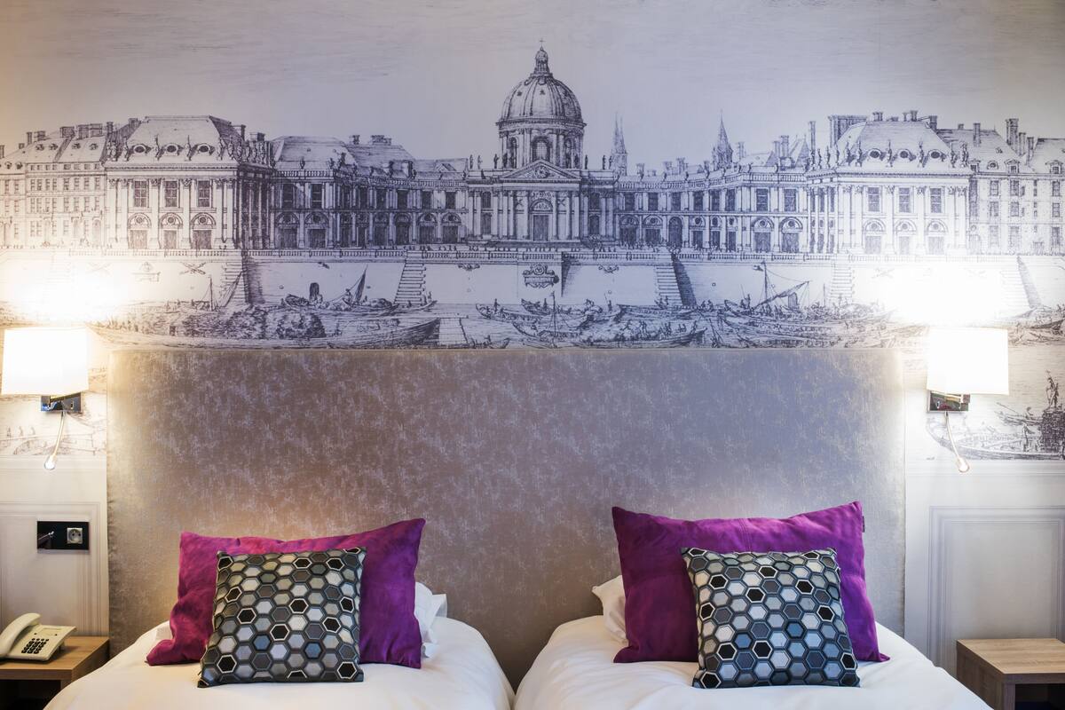 Una habitación de hotel con una cama grande, dos almohadas moradas, un cabecero con un mural de un paisaje urbano y una mesita de noche con una lámpara y un teléfono.