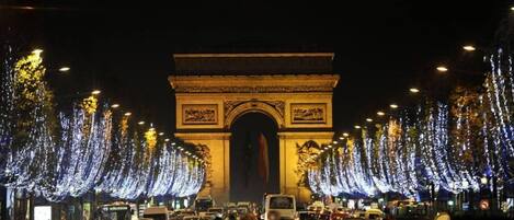 A nighttime view of the Arc de Triomphe with illuminated trees and a busy street.