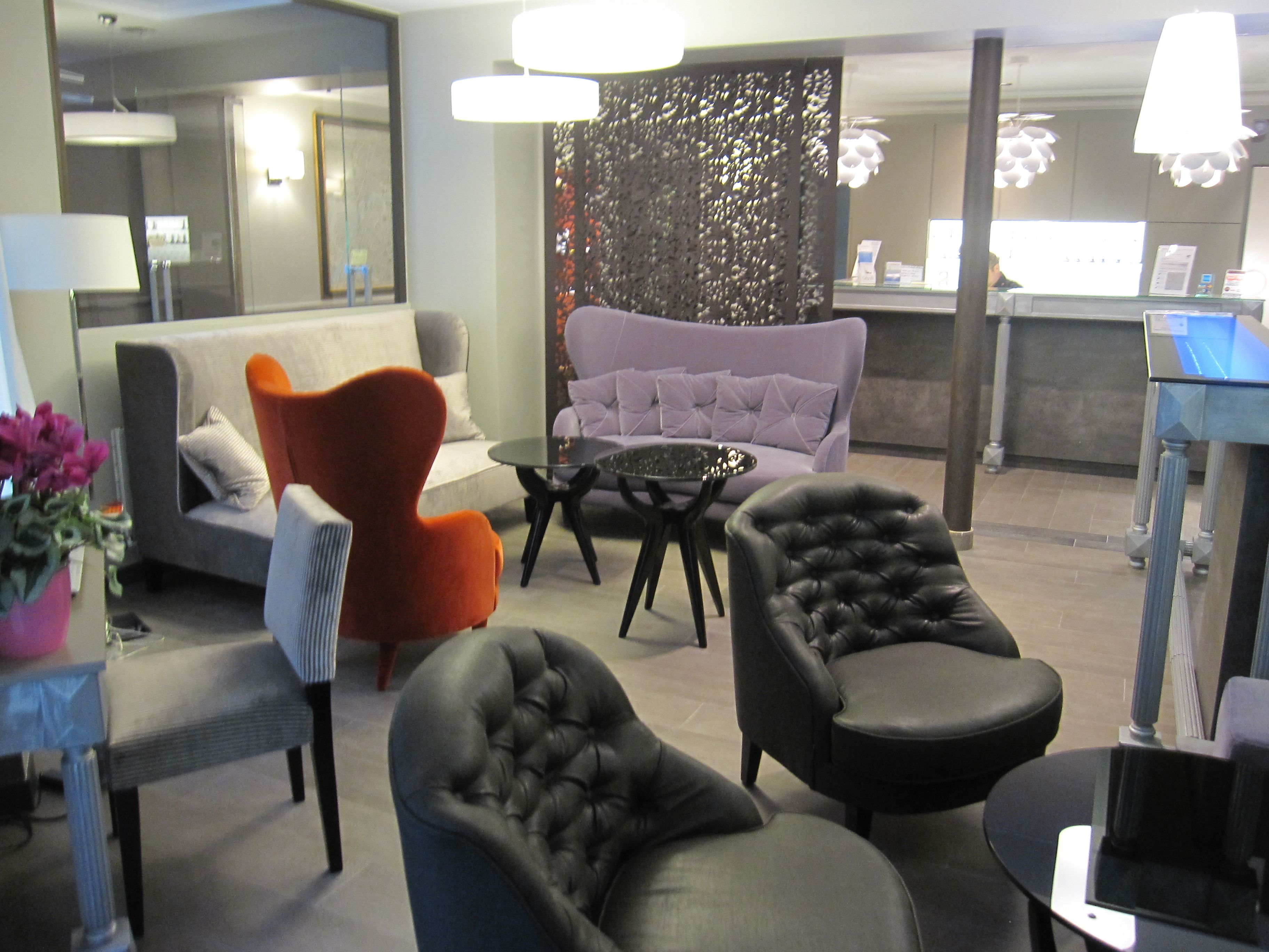 Et moderne loungeområde med en grå sofa, en orange lænestol og to sorte lænestole. Et sofabord i glas står i midten.