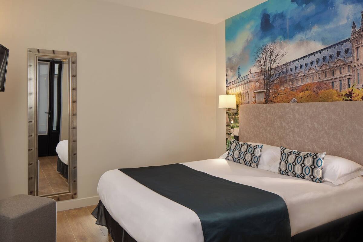 Habitación de hotel con una cama grande, un espejo, un cuadro de un edificio y una lámpara.
