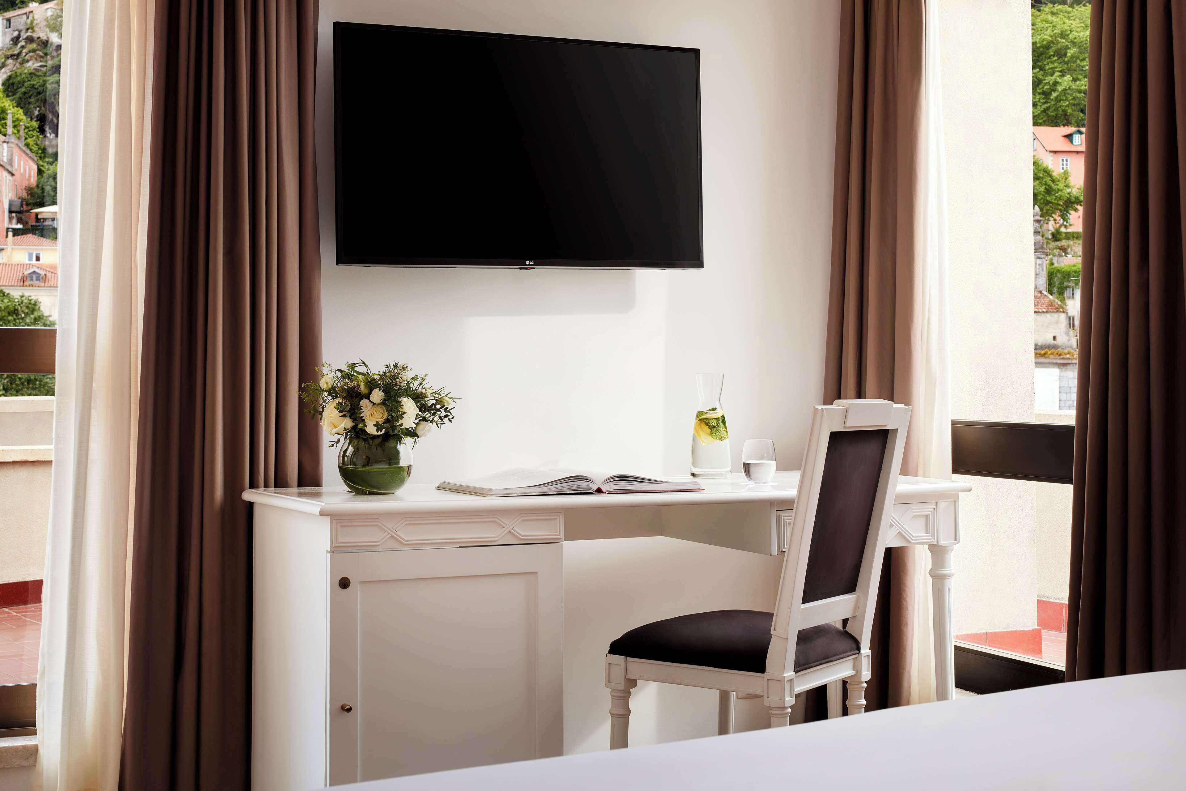 A hotel room with a white desk, a flat-screen TV mounted on the wall, a chair, a vase with flowers, and a book.