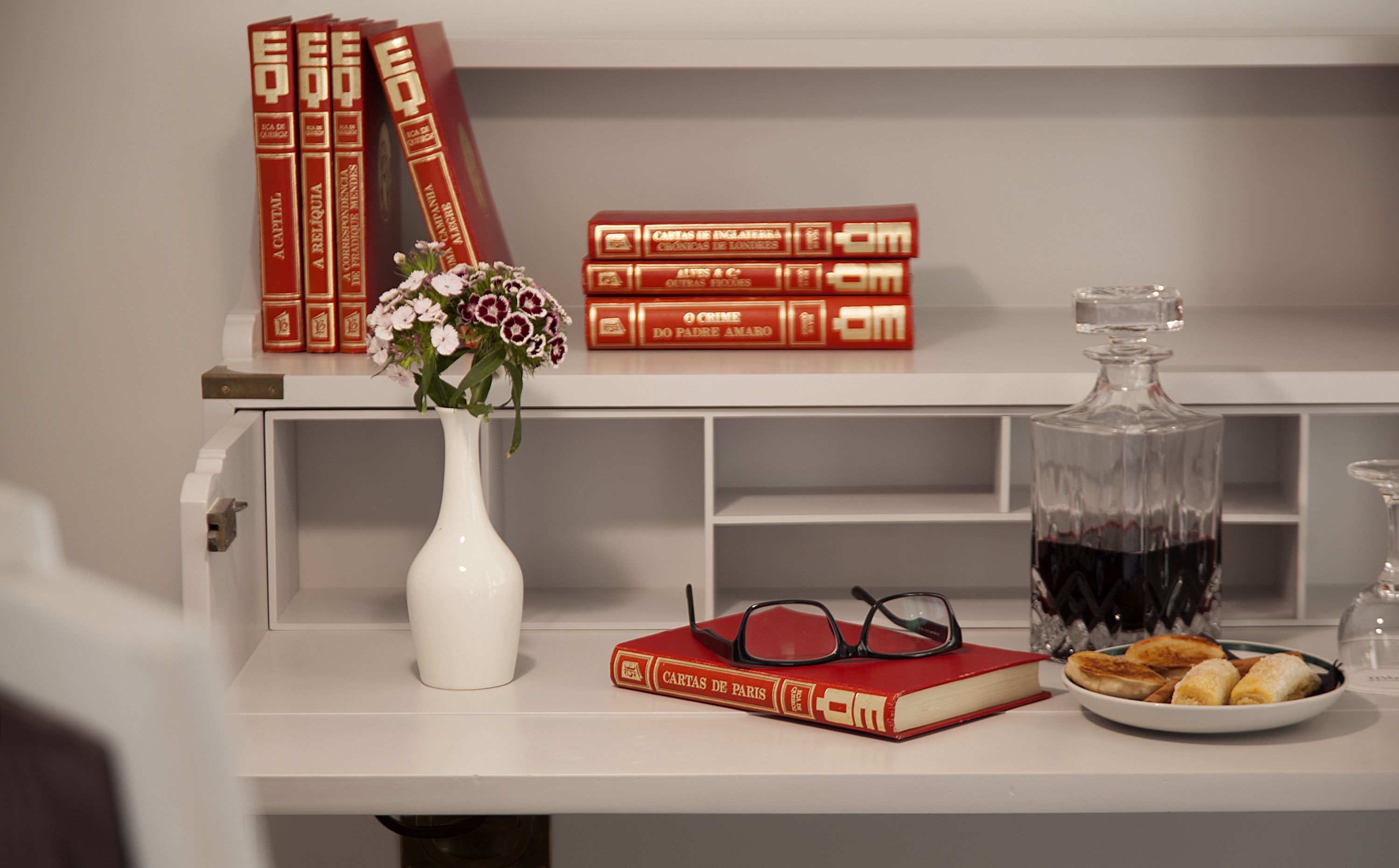 Una estantería con libros, un florero con flores, una botella de vino, copas y un plato de pasteles.
