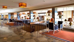 Un hall espacioso con suelo a cuadros, una zona de bar central con asientos y ventanas amplias que dejan entrar luz natural.