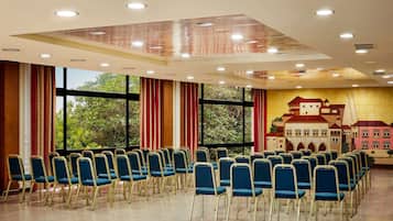 Un salón de conferencias con filas de sillas azules, ventanas amplias con cortinas rojas y un mural de un edificio en la pared.