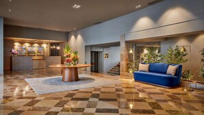 Un vestíbulo con piso a cuadros, una mesa de madera redonda con un centro de mesa floral, un sofá azul con cojines color beige y un mostrador de recepción con un fondo de paisaje urbano.