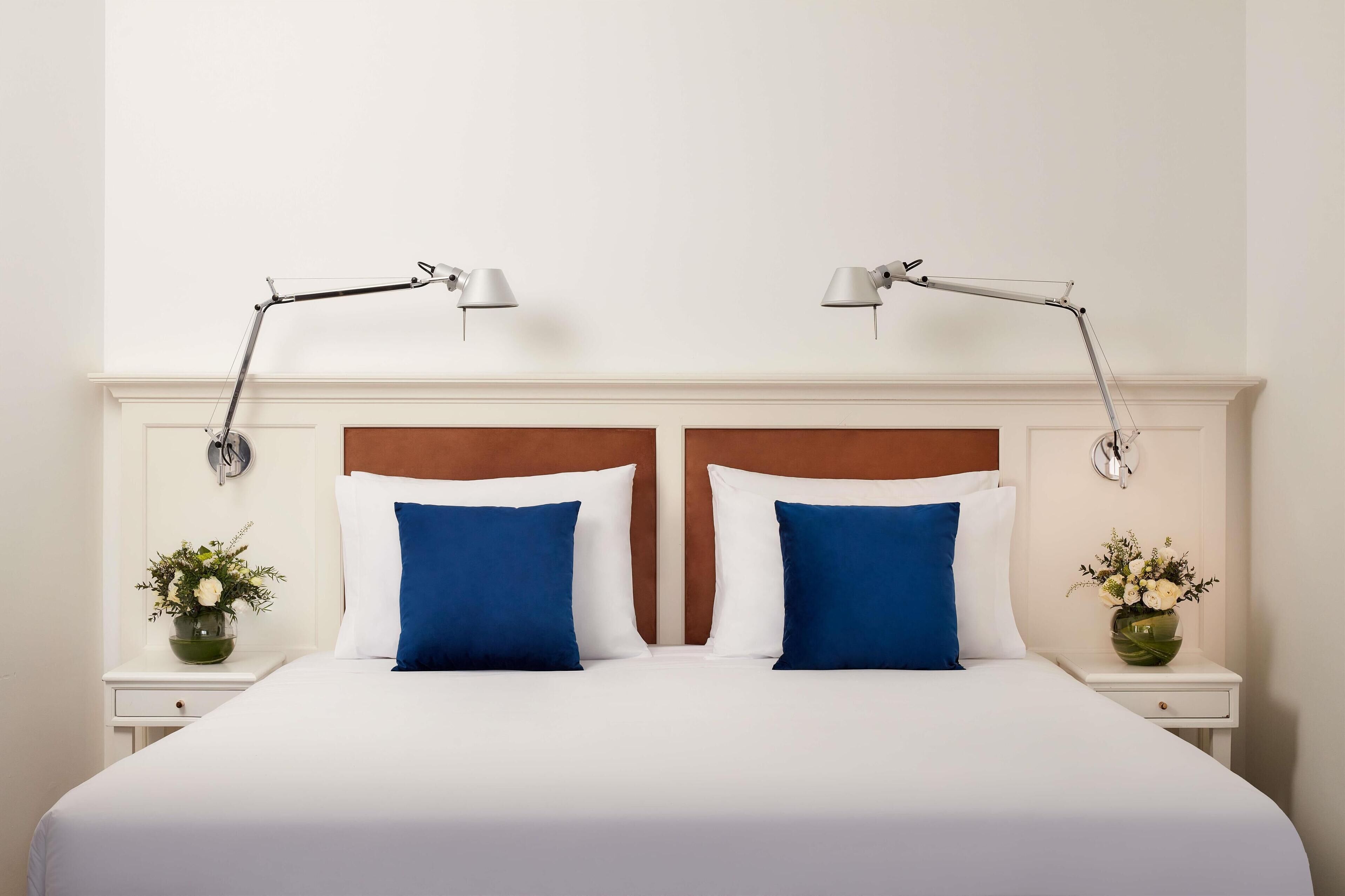 Un lit bien fait, avec des oreillers blancs et bleus, deux tables de chevet avec des lampes, et un vase de fleurs de chaque côté.