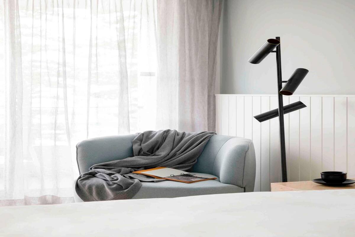 A room with a light blue armchair, a black floor lamp, a white radiator, and a window with sheer curtains.