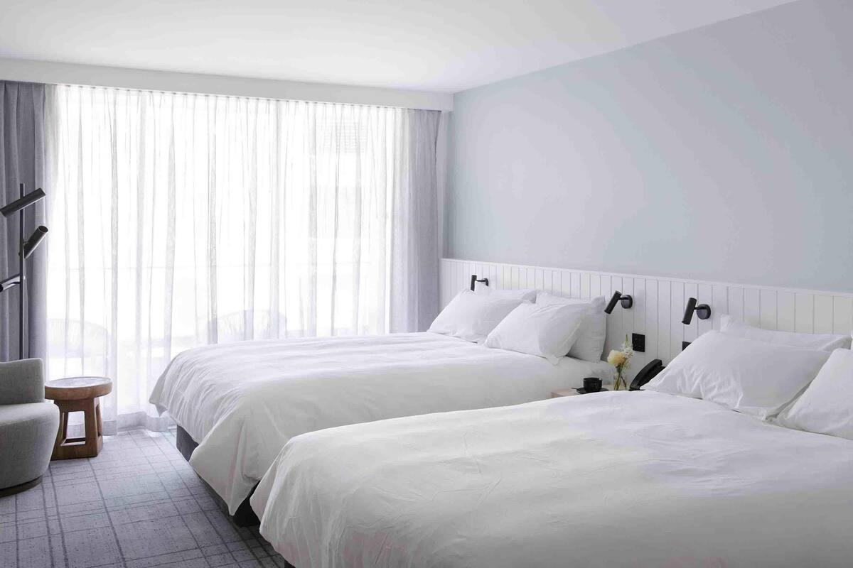 A hotel room with two beds, a small round table, and a large window with sheer curtains.