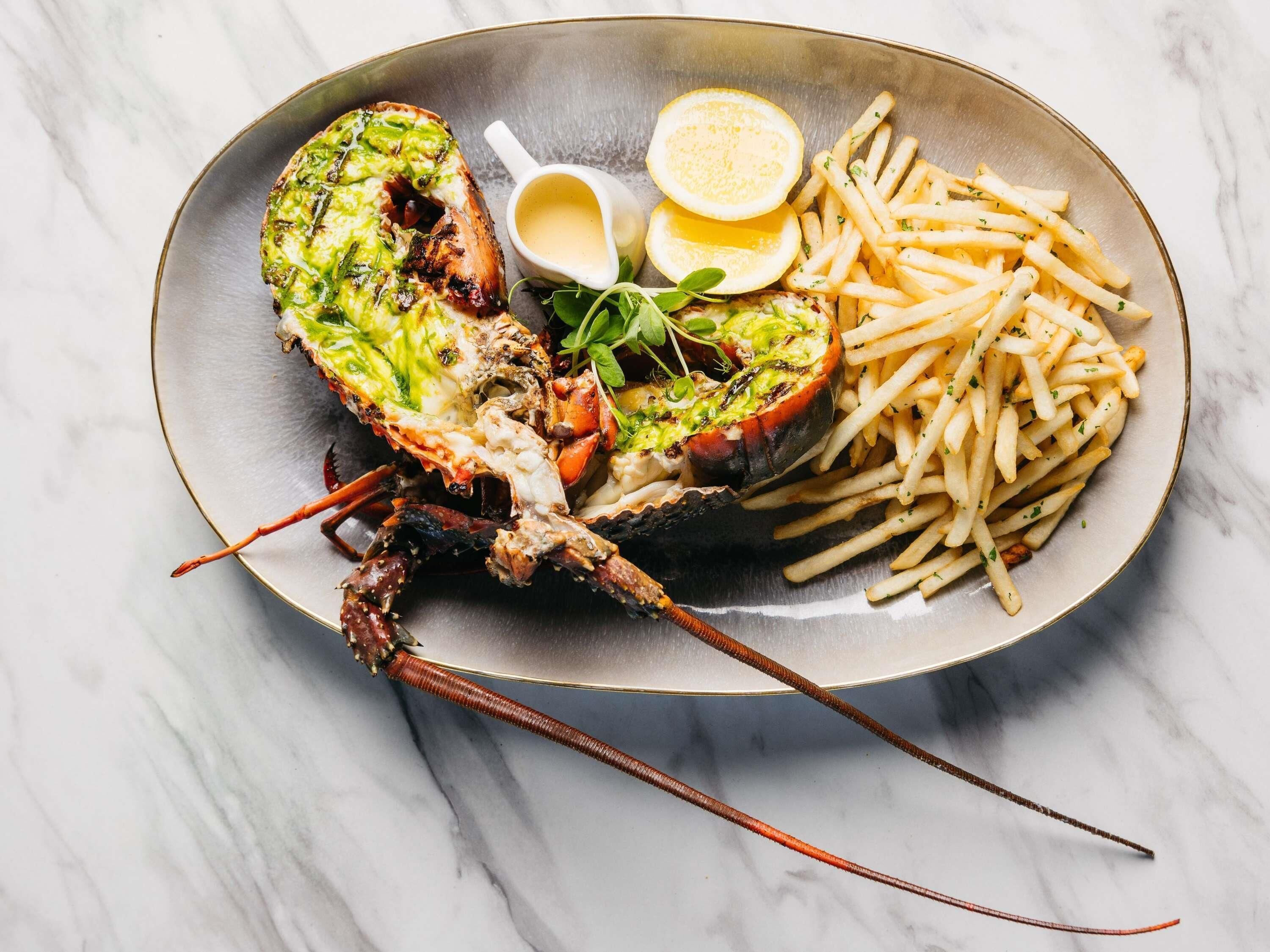 A lobster tail with a green topping, served with fries and lemon wedges on a plate.