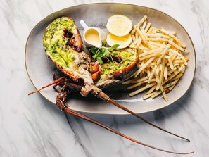 A lobster tail with a green topping, served with fries and lemon wedges on a plate.