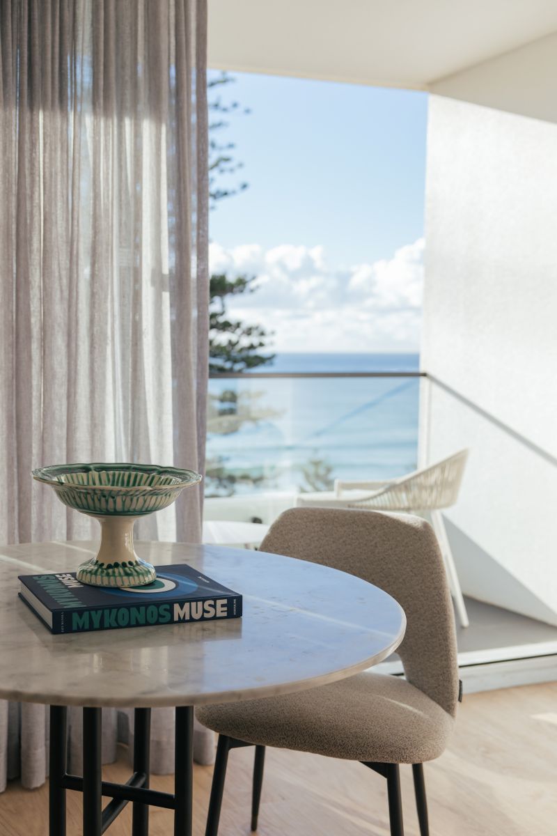 A modern room with a round table, a book titled 'MYKONOS MUSE,' and a view of the sea through a balcony.