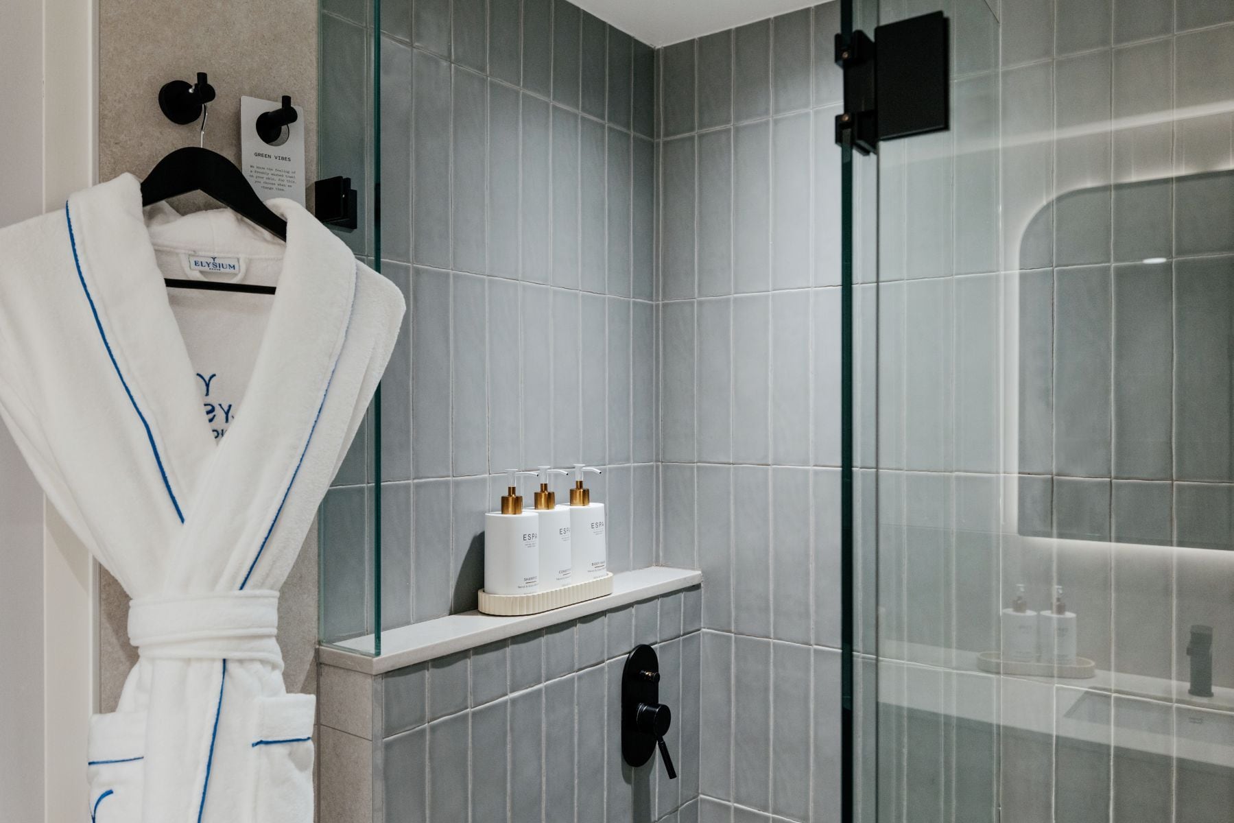 Un baño moderno con baldosas grises, cabina de ducha de vidrio, una bata blanca colgada en un perchero y artículos de aseo en un estante.