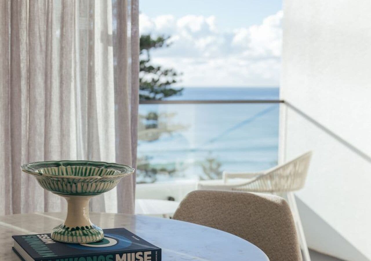 Uma mesa com uma tigela decorativa e um livro intitulado 'MUSE' numa varanda com vista para o mar.